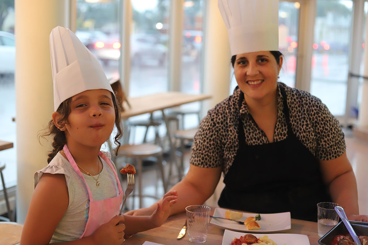 A mom and daughter in red aprons smiling with a plate of food at The Real Food Academy Miami, FL.